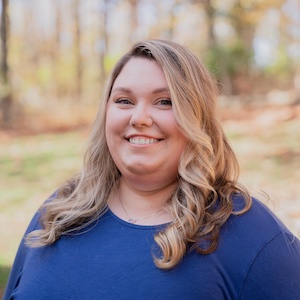 Photo of a woman with long blonde hair standing in front of a line of trees, blurred in the background.