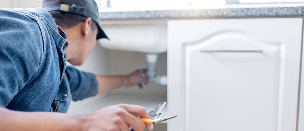 A man inspecting the underside of a kitchen sink while holding a clipboard.