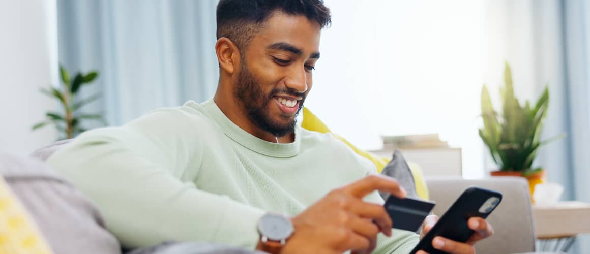 Young man checking credit on smartphone holding credit card and smiling on couch at home.