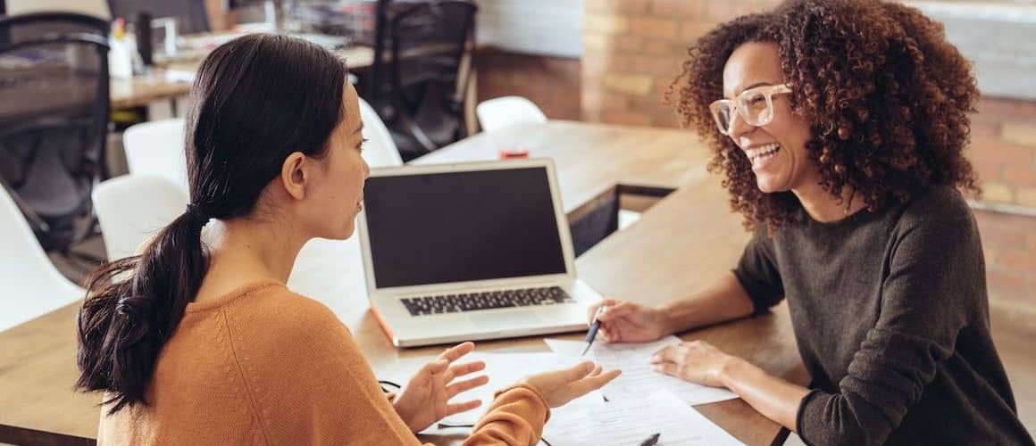 Two women discussing something over documents on a desk.