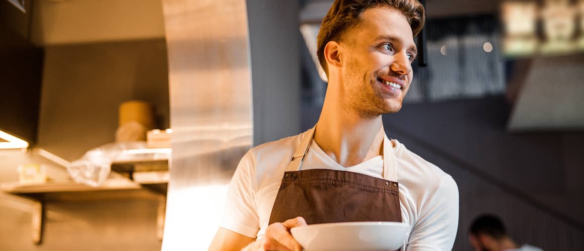 Waiter taking food from kitchen to serve at customer table, smiling.