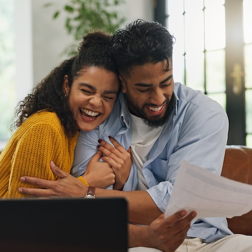 Young couple sitting on couch in sunny living room reading over their mortgage preapproval paperwork.
