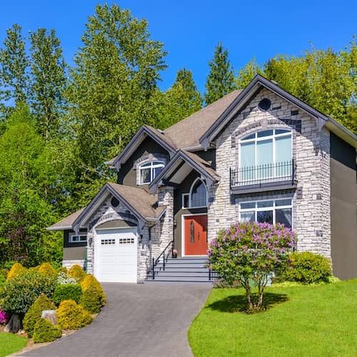 Large green sided and stone home surrounded by lush landscaping and mature trees.