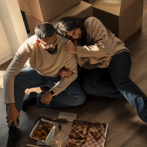 Couple snuggling and eating pizza on floor of new home surrounded by unopened moving boxes.