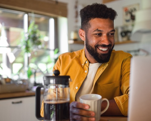 Man smiling, drinking coffee and looking at open laptop in kitchen.
