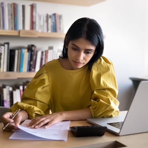 Young woman reviewing paperwork with calculator and laptop.