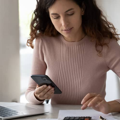 A young woman sitting at a desk, possibly managing bills or finances using a digital tablet.