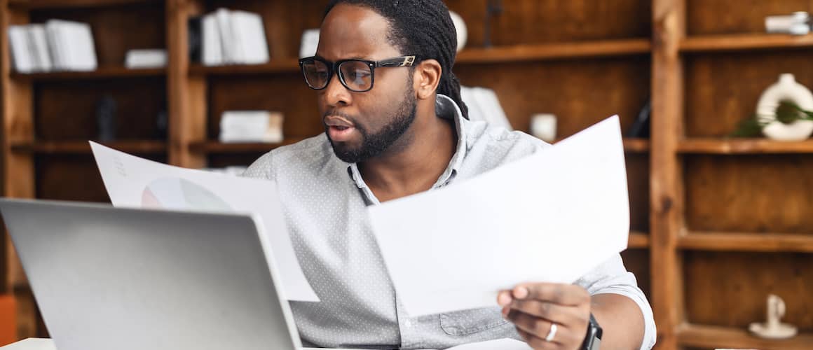 African American man reviewing paperwork in front of his laptop at his desk.