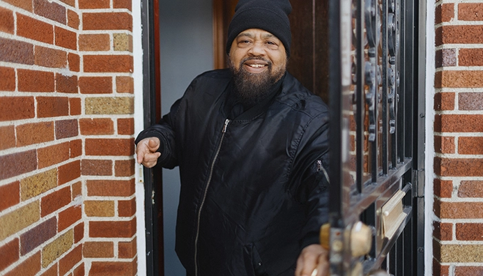 Man in hat smiling while opening front door on brick house.