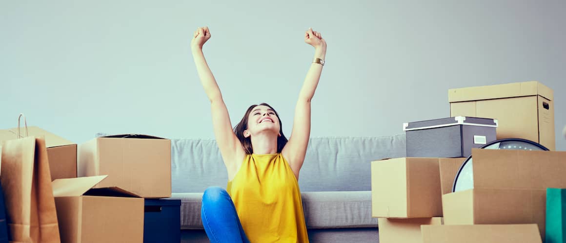 Happy woman with arms stretched overhead in joy, surrounded by moving boxes and in front of a grey couch.