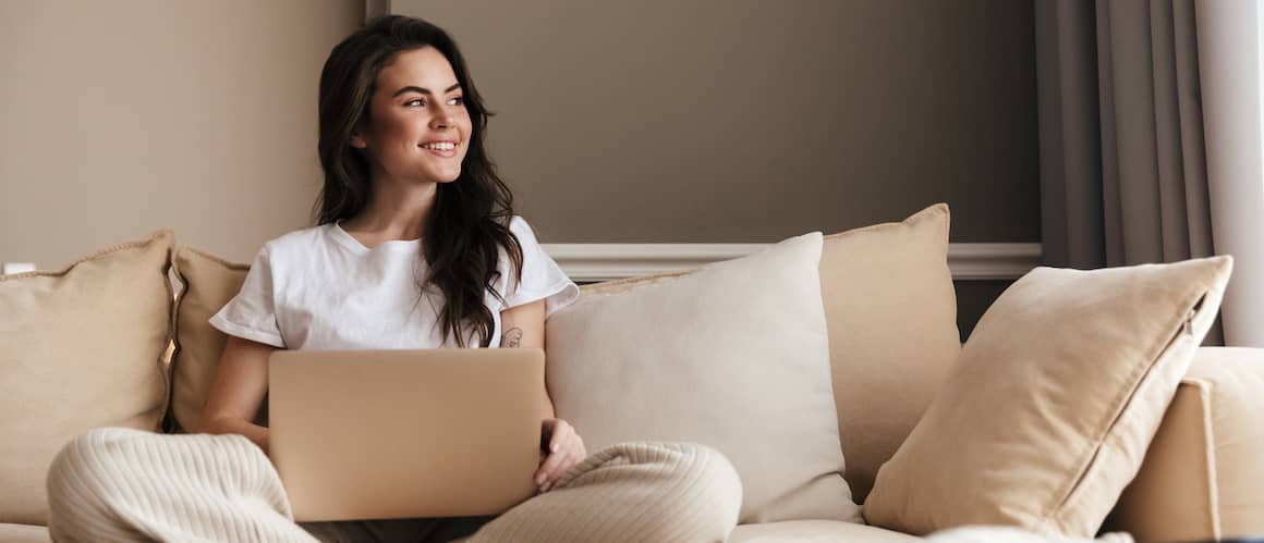 Young woman sitting on tan couch working on laptop and smiling.