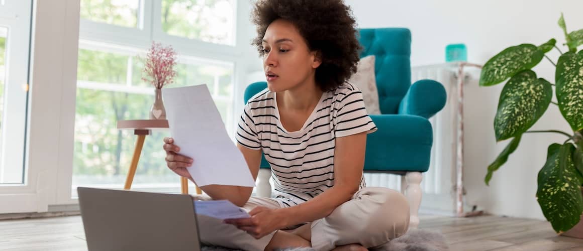 Young woman sitting cross legged on the floor in front of an open laptop while looking at a sheet of paper.