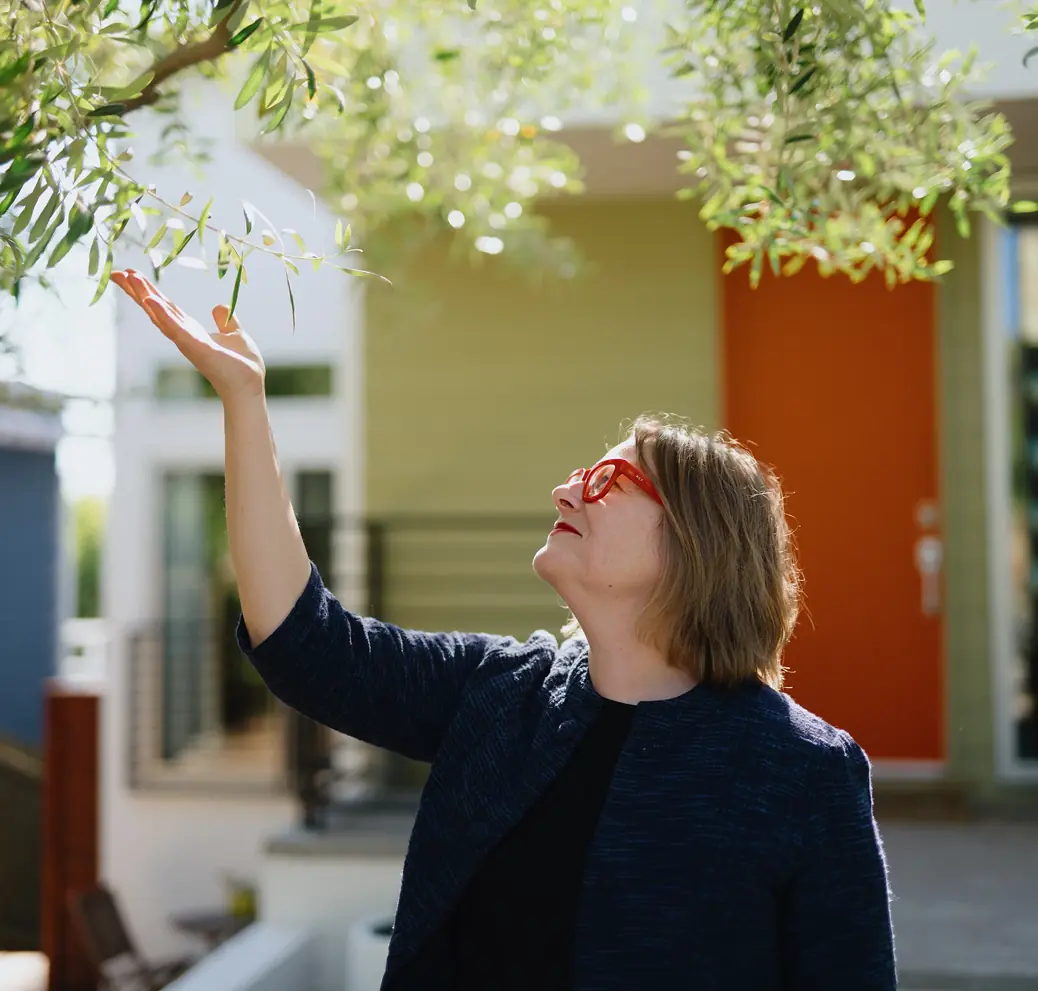 Woman reaching toward a leafy tree in a sunny courtyard.