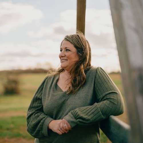 Woman wearing green long sleeve top leaning on wooden railing looking off into the distance.