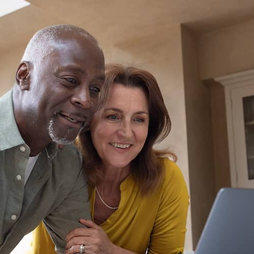 Retired couple in their kitchen looking at open laptop together and smiling.
