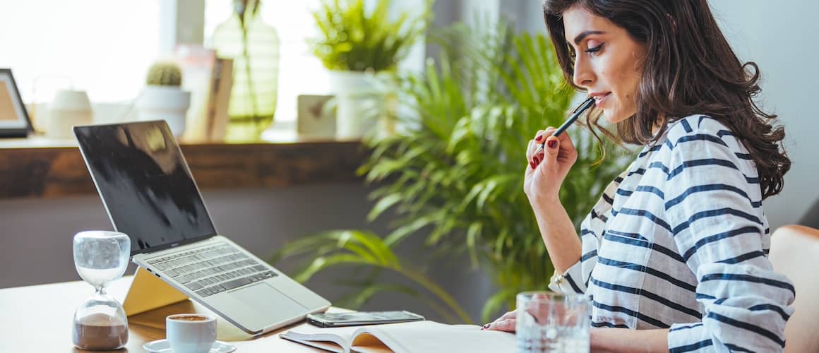 Woman at home office desk looking at notebook with open laptop and coffee nearby.