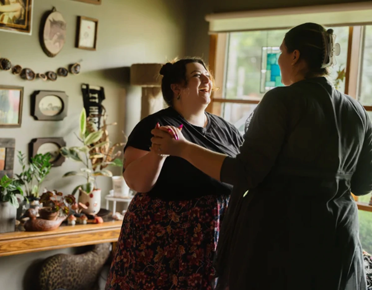 Two women dance together inside a cozy, plant-filled room. One smiles up at the other as they hold hands near a window with stained glass, surrounded by framed art and warm natural light.