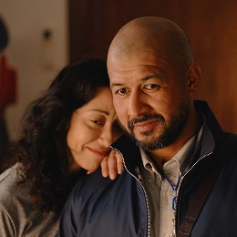 A woman rests her head on a man’s shoulder, smiling gently, while he looks ahead with a calm expression. They stand close together indoors under warm lighting, conveying affection and comfort.