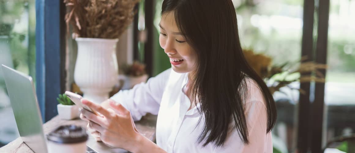 Young woman sitting down at a cafe in front of her laptop while using her phone.