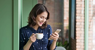 Una mujer blanca de pie afuera con una taza de café, mirando su teléfono. 