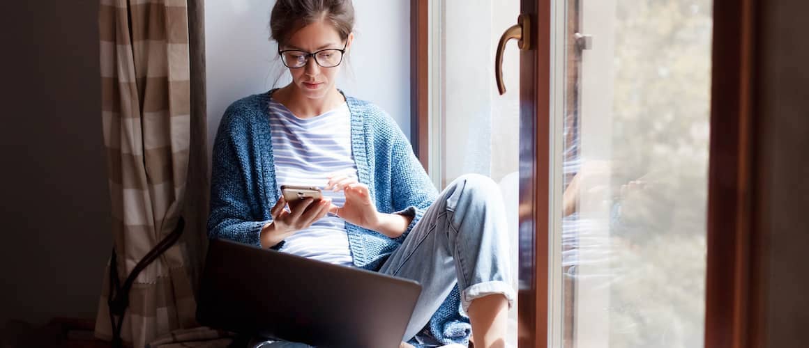 Woman checking credit score via phone on home window seat.