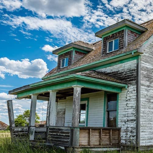 Image of distressed grey ranch-style family house with an unkempt overgrown lawn in disrepair.