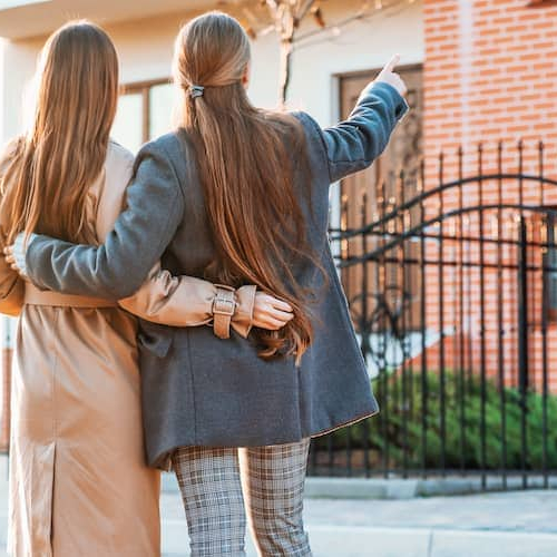 The image shows young female couple pointing at a home, possibly considering it for purchase.