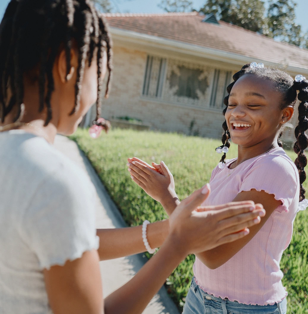 Two young Black girls in the driveway of their home clap their hands while playing a game together.