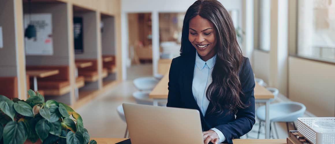 An African American businesswoman smiling while using her laptop at a table, likely in a professional setting.