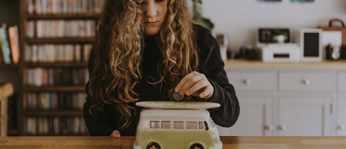 Girl with long hair putting money into a piggy bank shaped like a bus.