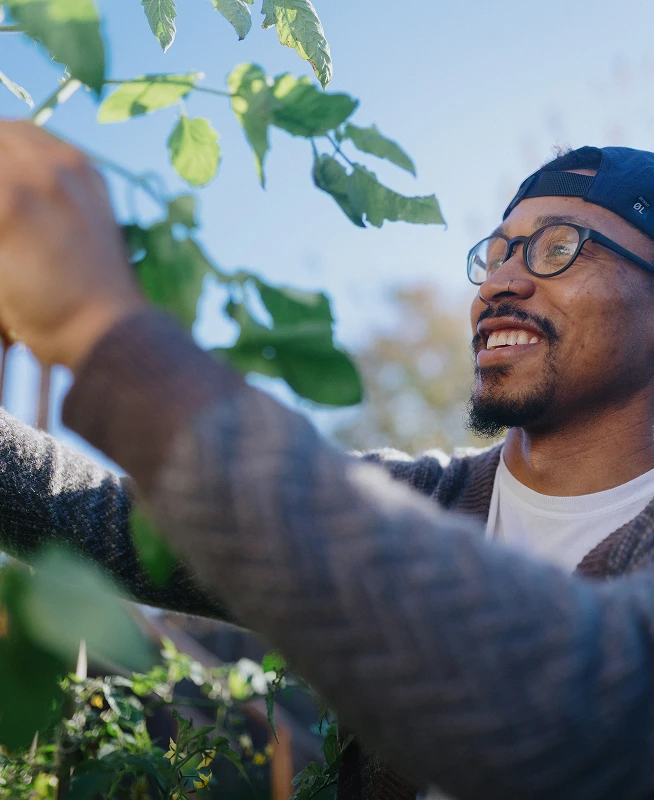 Black man with glasses smiles while reaching up to tend a tall garden plant