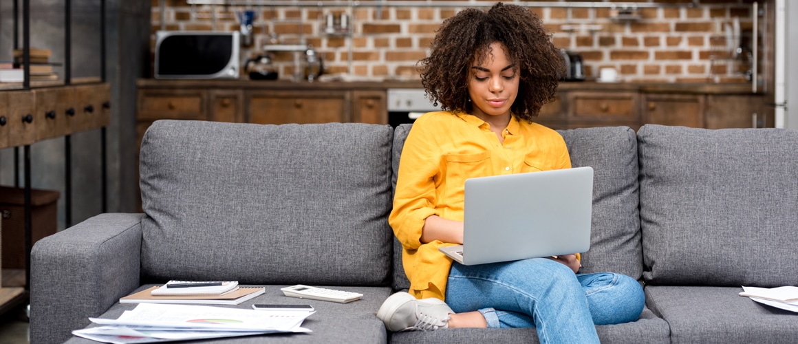 A women engrossed in her work on laptop seated on a couch with a bunch of papers lying near.