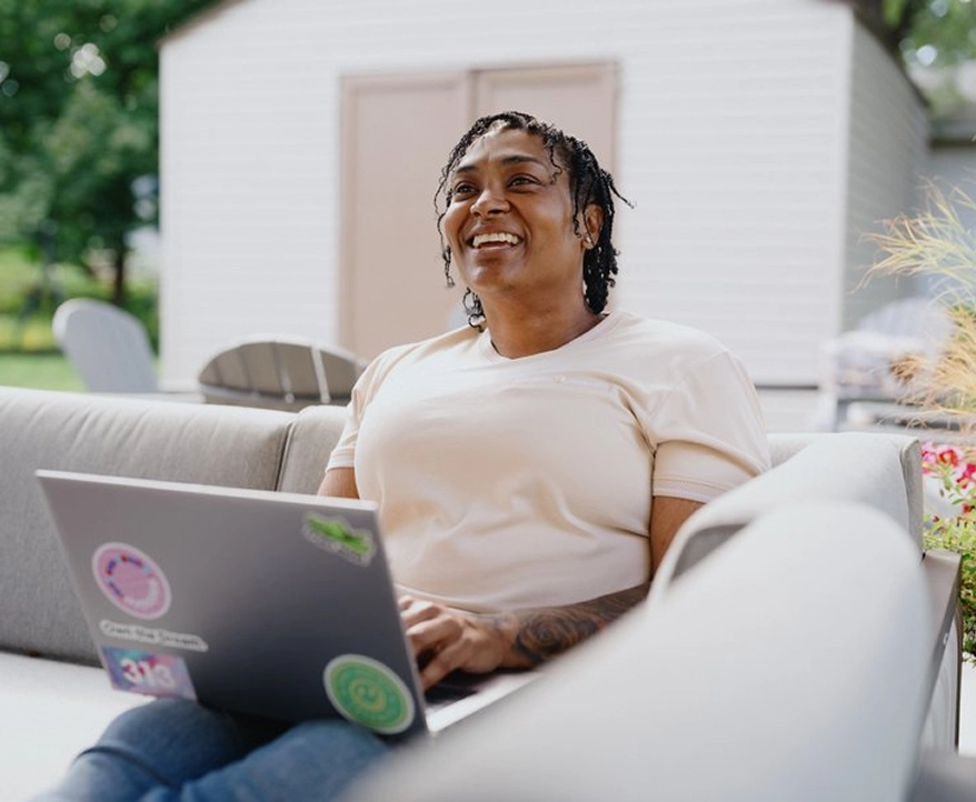 A woman wearing a light yellow shirt, sitting outdoors on a sofa, working on a laptop looking upward appearing relaxed and happy.