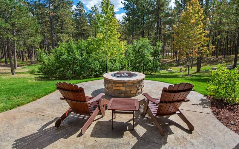 Two brown Adirondack chairs surrounding a stone fire pit and overlooking a beautiful woodsy backyard.