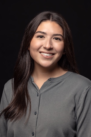 Headshot of a woman with long dark hair smiling in front of a black background.