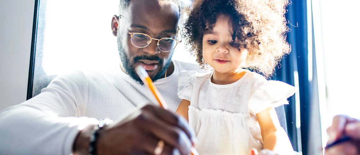 An image capturing a father and his child huddled closely together, both focused intently on a piece of paper laid out before them. The father, pencil in hand, is writing on the paper, while the child watches with interest.