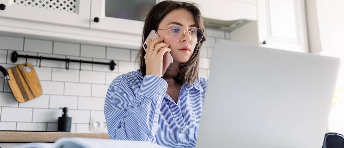 A woman sitting at the kitchen table looking at her computer and on the phone.