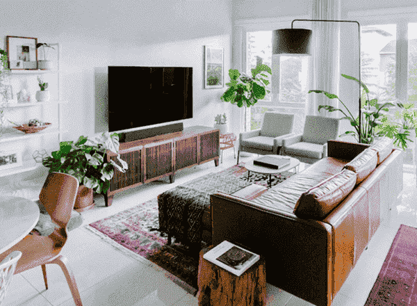 Open floor plan dining and living space with both boho and midcentury decor and houseplants placed throughout. Floating leather couch faces tv and white minimalist dining room table and oriental rug anchor the room.