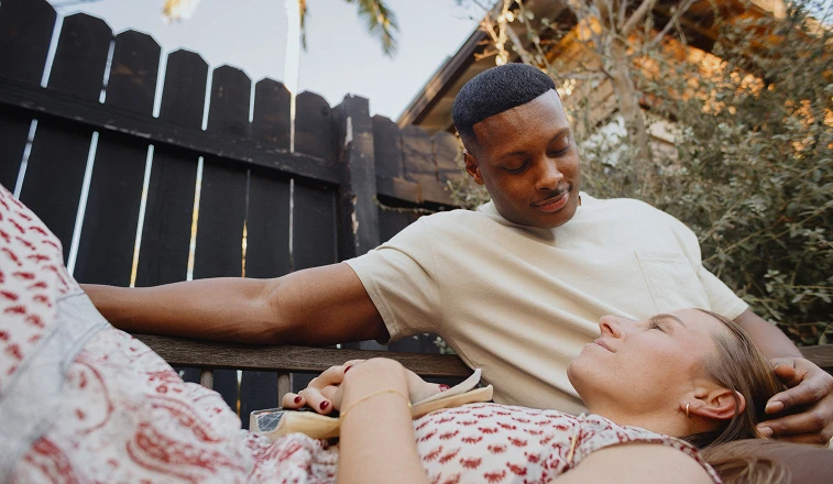 A man on a backyard bench with his partner resting her head on his lap.