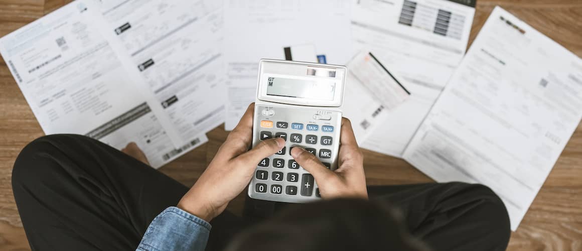 Top down view of a man sitting cross legged on the floor, with bills and a calculator, figuring out his budget.