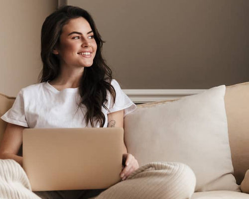 Young woman sitting on tan couch working on laptop and smiling.