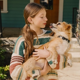 A young girl wearing a striped sweater holds a small tan dog in her arms, looking at the dog as it gazes back at her.