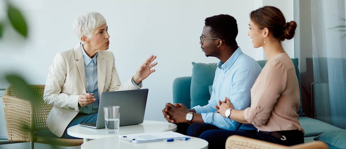 Financial advisor speaking with a young couple in an office with a laptop open on a table.