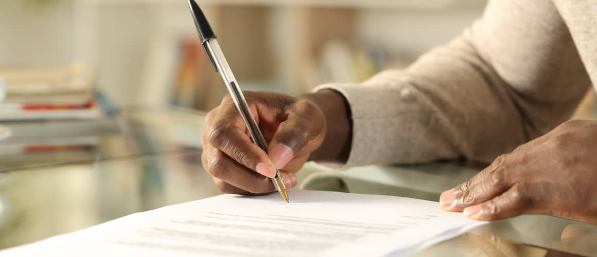 A Black man's hands signing a document on a desk at home, potentially related to paperwork or contracts.