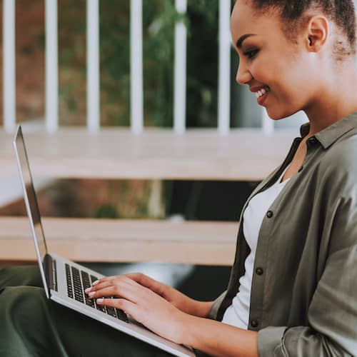 Black woman on patio on laptop smiling, showing a woman on a patio, potentially working on a laptop and smiling.