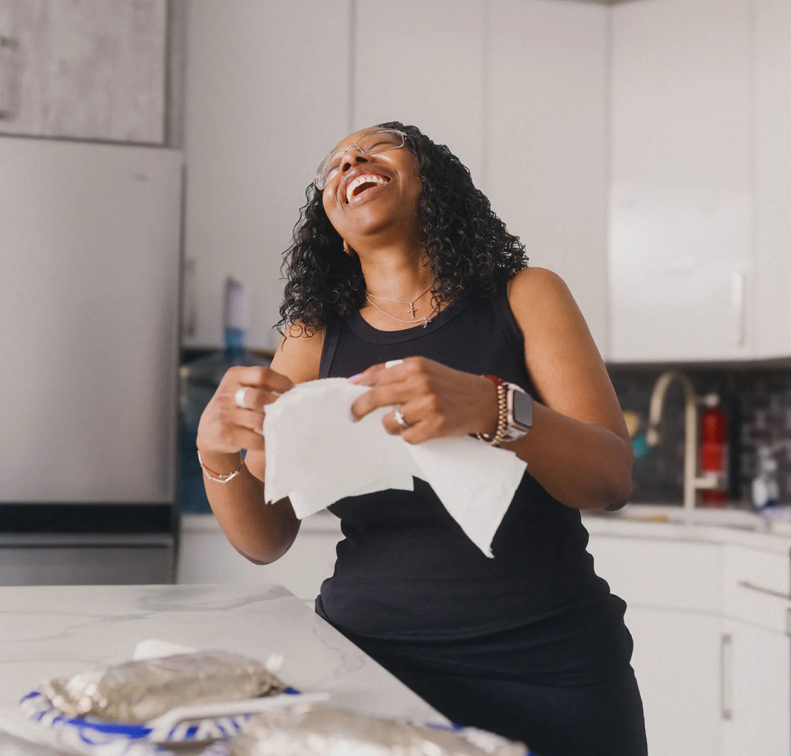 A Black woman stands in a kitchen with white cupboards, holding paper napkins while she throws her head back and laughs joyfully.