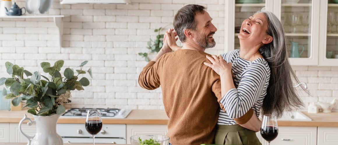 Couple dancing in kitchen with veggies, wine, and other food on the counter.