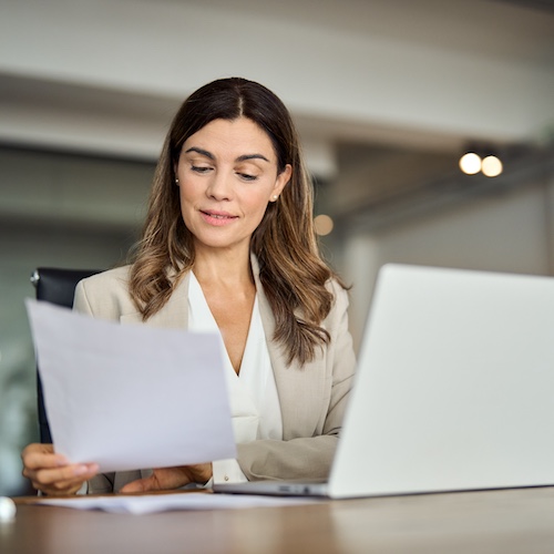 Young woman looking at papers in her hand while having a laptop in front of her.