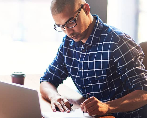 Focused man at desk writing down notes while desktop and laptop.
