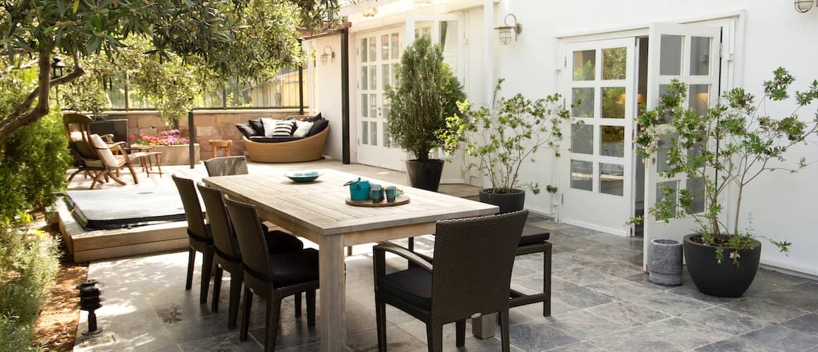 Backyard patio with wooden table and brown chairs with many plants around them.